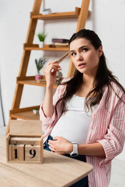 Selektiver Fokus der nachdenklichen und schwangeren Frau, die Haare in der Nähe von Holzwürfeln mit Datum berührt — Stockfoto