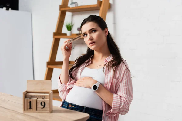 Selektiver Fokus der nachdenklichen und schwangeren Frau, die den Bauch in der Nähe von Holzwürfeln mit Datum berührt — Stockfoto