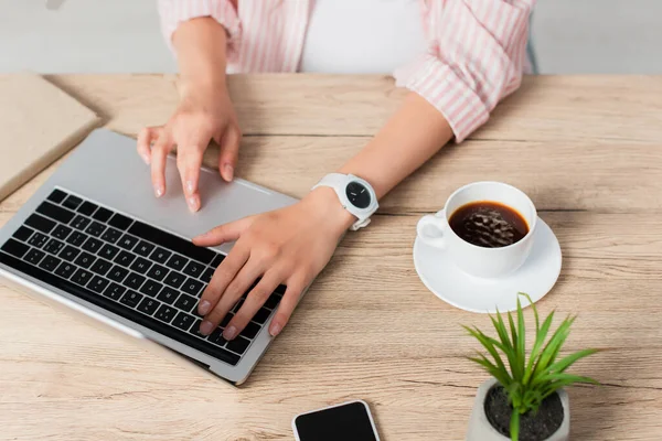 Partial view of freelancer using laptop near cup of coffee, plant and smartphone with blank screen — Stock Photo