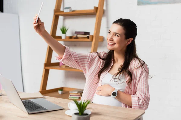 Selective focus of pregnant freelancer taking selfie near laptop — Stock Photo