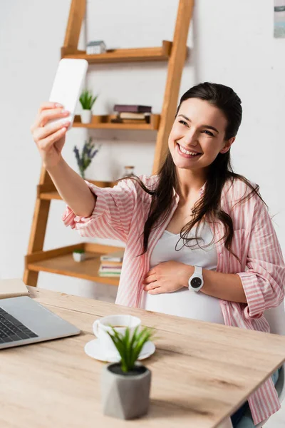 Messa a fuoco selettiva della donna incinta prendendo selfie vicino al computer portatile — Foto stock