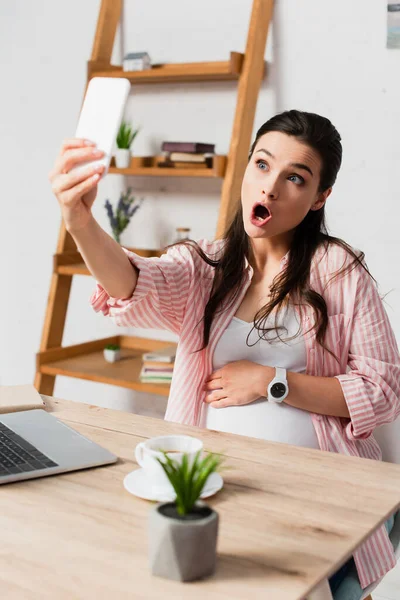 Enfoque selectivo de la mujer sorprendida y embarazada tomando selfie cerca de la computadora portátil - foto de stock