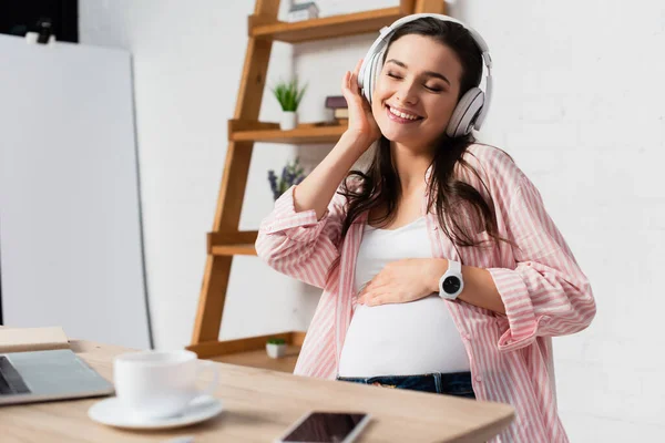 Enfoque selectivo de la mujer embarazada con los ojos cerrados escuchando música en auriculares inalámbricos cerca de gadgets y taza - foto de stock