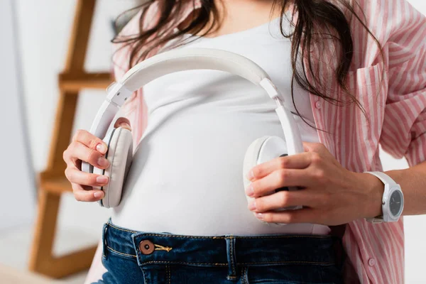Vue recadrée de la femme enceinte tenant des écouteurs sans fil près du ventre — Photo de stock