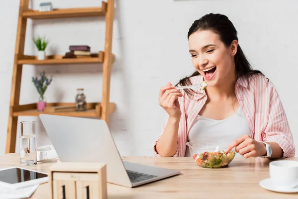 Brünette Frau blickt auf Laptop neben Salat, digitales Tablet und Tasse auf Tisch — Stockfoto