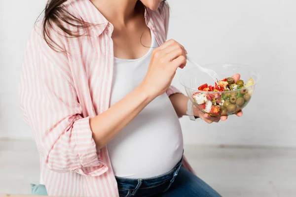 Vista parcial de la mujer embarazada sosteniendo tenedor de plástico cerca de contenedor para llevar con ensalada fresca - foto de stock