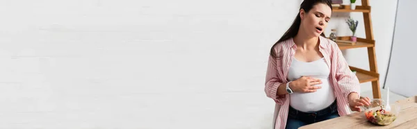 Panoramic shot of pregnant woman touching belly while suffering from pain near takeaway container with fresh salad — Stock Photo