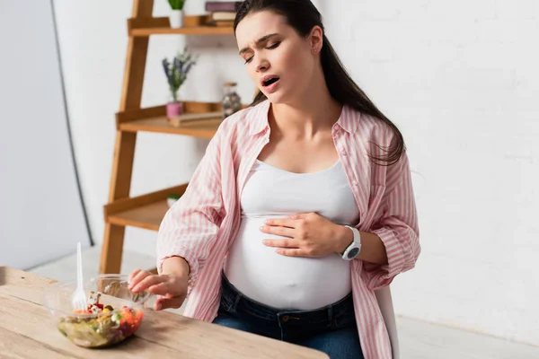 Беременная женщина трогает живот во время боли и сидит рядом с контейнером для еды со свежим салатом — стоковое фото