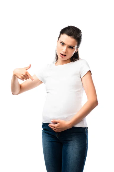 Mujer embarazada joven en camiseta blanca apuntando con el dedo hacia el vientre aislado en blanco - foto de stock