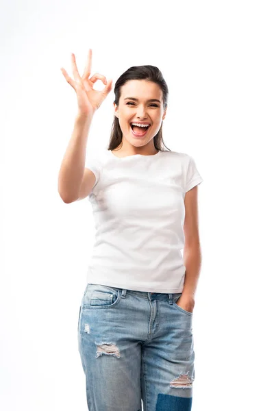 Молода жінка в білій футболці і джинсах стоїть з рукою в кишені і показує знак ОК ізольовано на білому — стокове фото