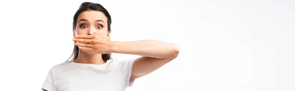 Tiro panorâmico de mulher chocada e morena em branco t-shirt cobrindo boca isolada em branco — Fotografia de Stock