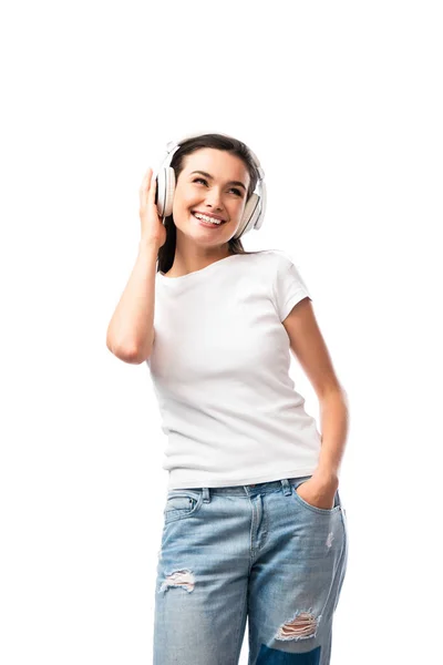 Mujer joven en camiseta blanca y auriculares inalámbricos de pie con la mano en el bolsillo aislado en blanco - foto de stock