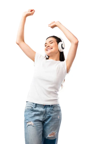 Mujer joven en camiseta blanca y auriculares inalámbricos bailando con las manos sobre la cabeza aisladas en blanco - foto de stock
