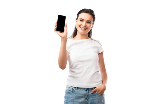 Молода жінка в білій футболці стоїть з рукою в кишені і тримає смартфон з порожнім екраном ізольовано на білому — стокове фото