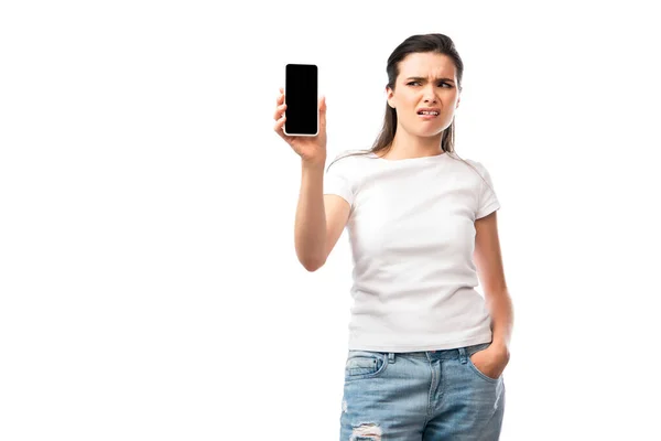 Mujer disgustada de pie con la mano en el bolsillo y mirando el teléfono inteligente con pantalla en blanco aislado en blanco — Stock Photo