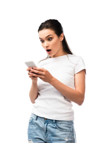 Шокированная брюнетка женщина в белой футболке с помощью смартфона изолированы на белом — стоковое фото