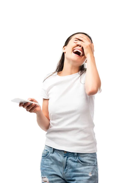 Молода жінка прикриває обличчя, сміється і тримає смартфон ізольовано на білому — стокове фото