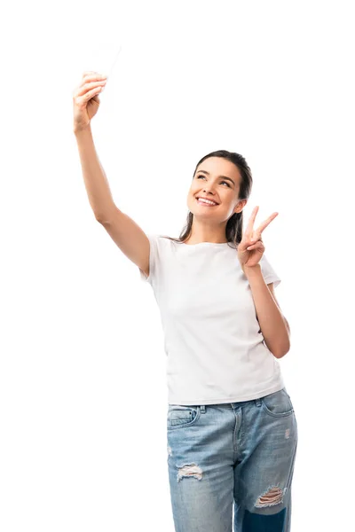 Брюнетка жінка в білій футболці бере селфі і показує знак миру ізольовано на білому — стокове фото