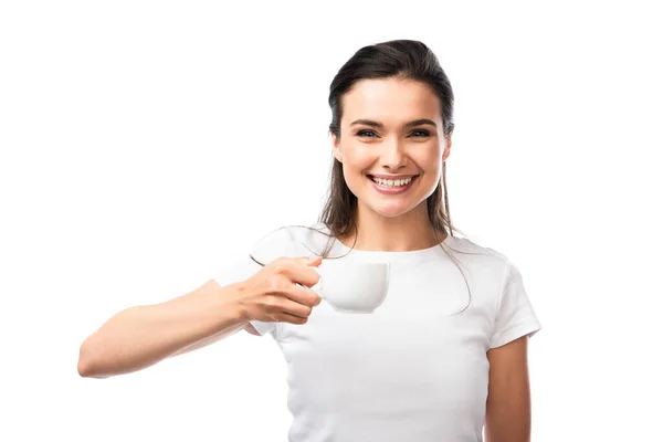 Morena mujer en camiseta blanca mirando a la cámara y sosteniendo taza con café aislado en blanco - foto de stock