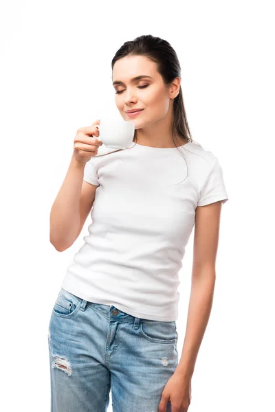 Молода жінка з закритими очима в білій футболці пахне кавою в чашці ізольовано на білому — стокове фото