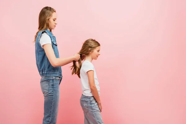 Side view of young woman in denim clothes braiding hair of daughter isolated on pink — Stock Photo