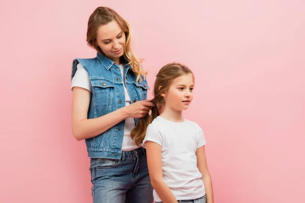 Молодая женщина в джинсовом жилете и джинсах плетение волос дочери изолированы на розовый — стоковое фото