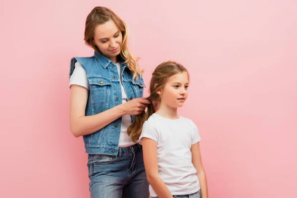 Mujer joven en chaleco vaquero y jeans trenzando pelo de hija aislada en rosa - foto de stock