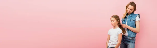 Панорамный снимок женщины в джинсовой одежде, плетущей волосы дочери, изолированной на розовом — стоковое фото