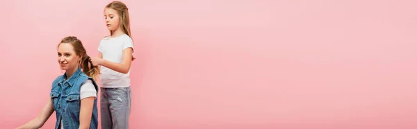 Imagen horizontal de niña en camiseta blanca y jeans trenzando pelo de madre aislada en rosa - foto de stock