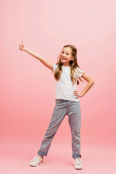 Bambino in t-shirt bianca con mano sul fianco mostrando pollice in su mentre guarda la fotocamera su rosa — Foto stock