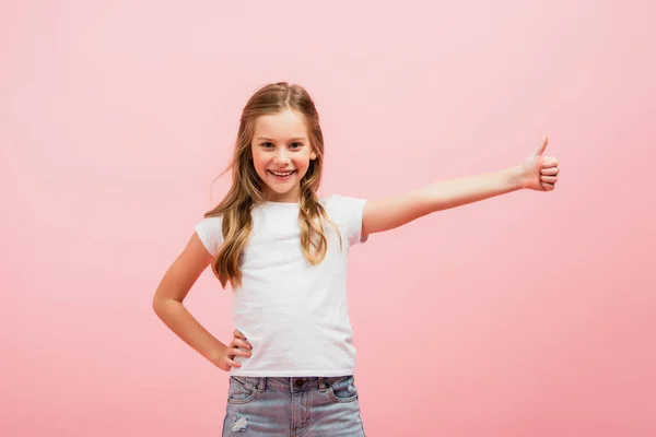 Bambino in t-shirt bianca con mano sul fianco mostrando pollice in su mentre guarda la fotocamera isolata su rosa — Foto stock