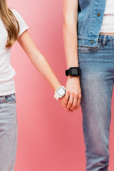 Vista recortada de la madre y la hija en jeans y smartwatches tomados de la mano en rosa - foto de stock