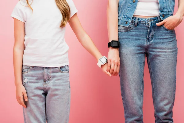 Visión parcial de la mujer y el niño en jeans y smartwatches tomados de la mano en rosa - foto de stock