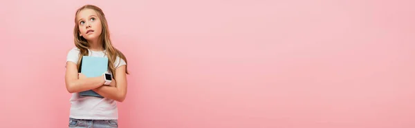 Conceito panorâmico de menina pensativa em smartwatch olhando embora enquanto segurando livro isolado em rosa — Fotografia de Stock