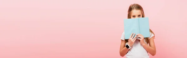 Imagen horizontal de chica en smartwatch oscurecer la cara con libro mientras mira la cámara aislada en rosa - foto de stock