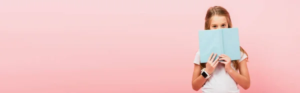 Horizontal image of girl in smartwatch obscuring face with book while looking at camera isolated on pink — Stock Photo