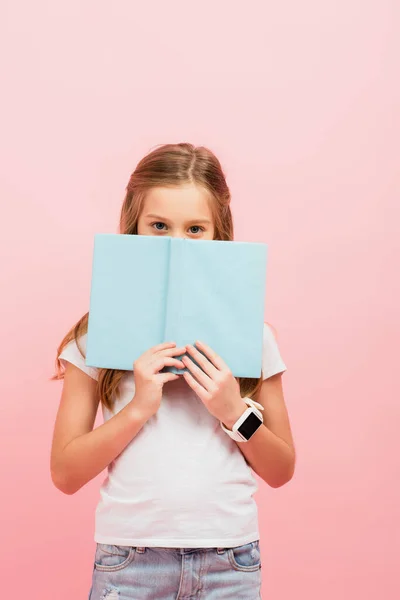 Ragazza in smartwatch volto oscuro con libro mentre guardando la fotocamera isolata su rosa — Foto stock