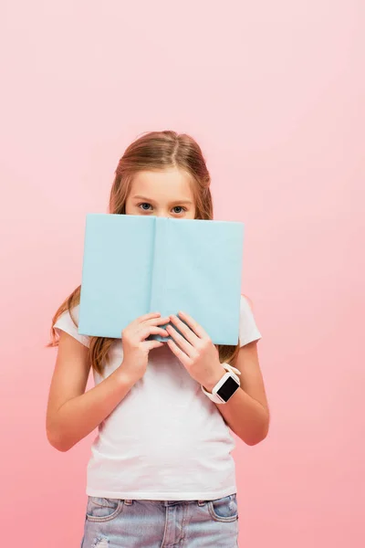 Chica en smartwatch oscurecer la cara con libro mientras mira la cámara aislada en rosa - foto de stock