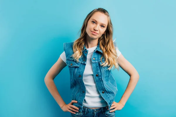 Junge Frau in Jeansweste steht mit den Händen auf den Hüften und blickt vereinzelt in die Kamera auf blauem Grund — Stockfoto