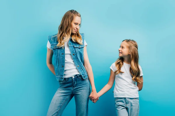 Criança em t-shirt branca de mãos dadas com a mãe em roupas jeans enquanto olham um para o outro isolado em azul — Fotografia de Stock