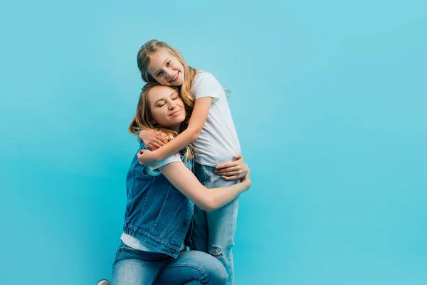 Madre con los ojos cerrados vistiendo ropa de mezclilla y abrazando hija aislada en azul - foto de stock