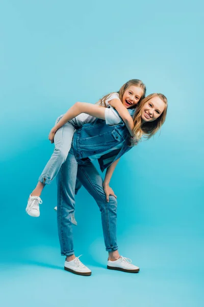 Full length view of young woman in denim vest and jeans piggybacking daughter while looking at camera on blue — Stock Photo