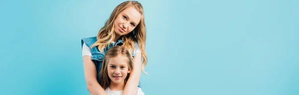 Horizontal image of young woman hugging daughter while looking at camera isolated on blue — Stock Photo