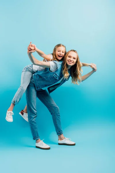 Piena lunghezza vista di giovane madre in denim vestiti piggybacking figlia mentre si tiene per mano su blu — Foto stock