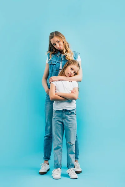 Full length view of upset woman in denim clothes hugging offended daughter in white t-shirt and jeans on blue — Stock Photo