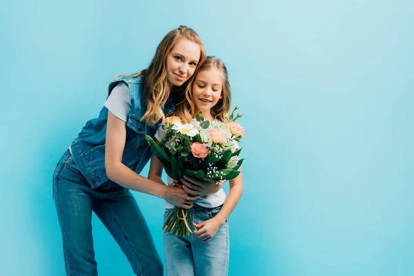 Jeune femme en denim présentant des fleurs à la fille tout en regardant la caméra isolée sur bleu — Photo de stock