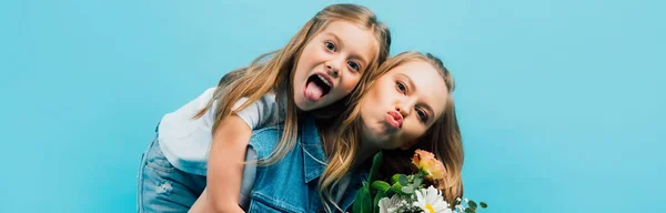 Colpo panoramico di ragazza che sporge la lingua e abbraccia le labbra della madre tenendo in mano mazzo di fiori isolati su blu — Foto stock