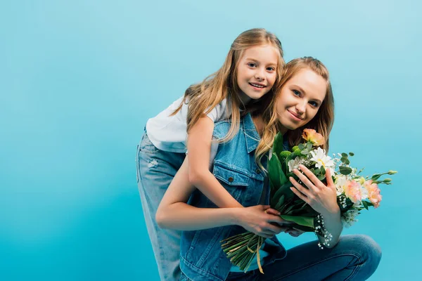 Child embracing young mother holding bouquet of flowers isolated on blue — Stock Photo