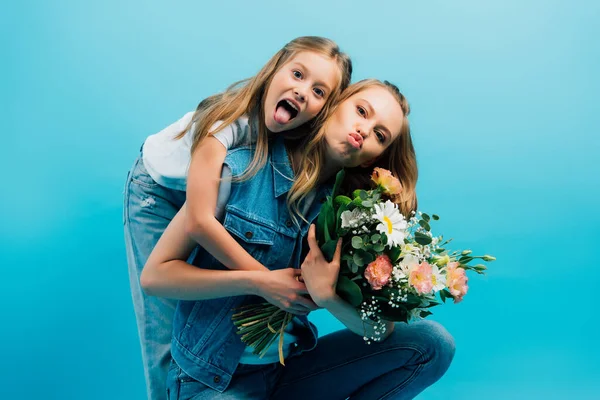 Fille qui sort la langue et étreint mère boudins lèvres tout en tenant bouquet de fleurs isolées sur bleu — Photo de stock