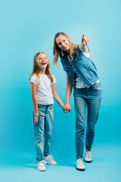 Vista completa de la chica emocionada cogida de la mano con la madre señalando con el dedo y mirando hacia otro lado en azul - foto de stock